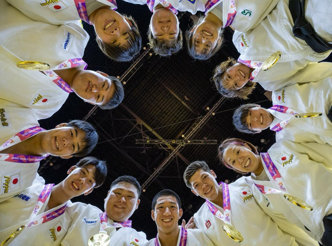 Judoka in white gis form a circle around the camera, smiling and holding gold medals.