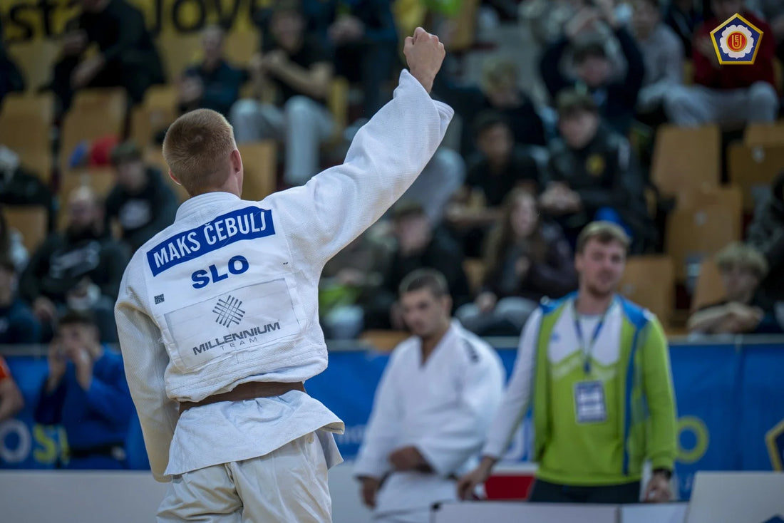 Judoka in a white gi raises a clenched fist toward the crowd, with a back patch reading Maks Čebulj and SLO.