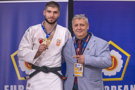 A judoka in a white gi holds up a medal beside a man wearing a light grey jacket and an accreditation badge.