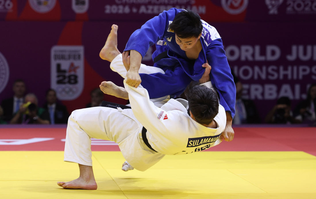 A judoka in a blue gi throws a judoka in a white gi above the yellow tatami during a contest.