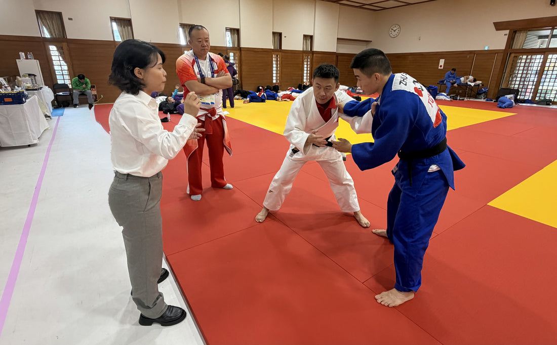 A woman gestures beside two judoka gripping each other on red and yellow tatami mats, with several people watching in an indoor dojo.