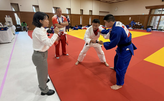 A woman gestures beside two judoka gripping each other on red and yellow tatami mats, with several people watching in an indoor dojo.