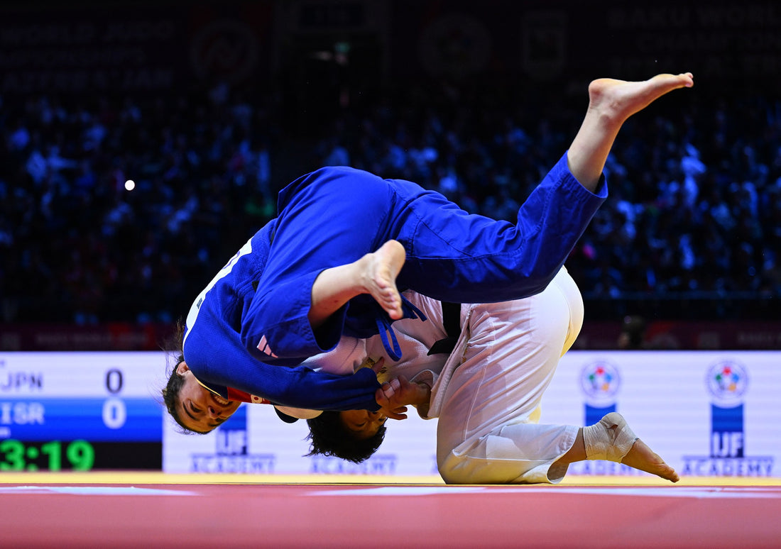 A judoka in a blue gi is airborne over an opponent in a white gi during a match on a red-and-yellow tatami.