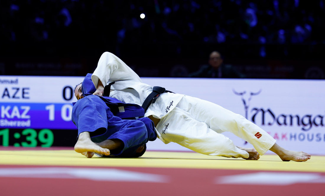 Two judoka in white and blue gis fight on the tatami as a scoreboard glows behind them.