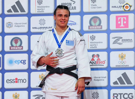 A judoka in a white judogi and black belt wears a medal and Greek flag patch while standing before a sponsor backdrop.