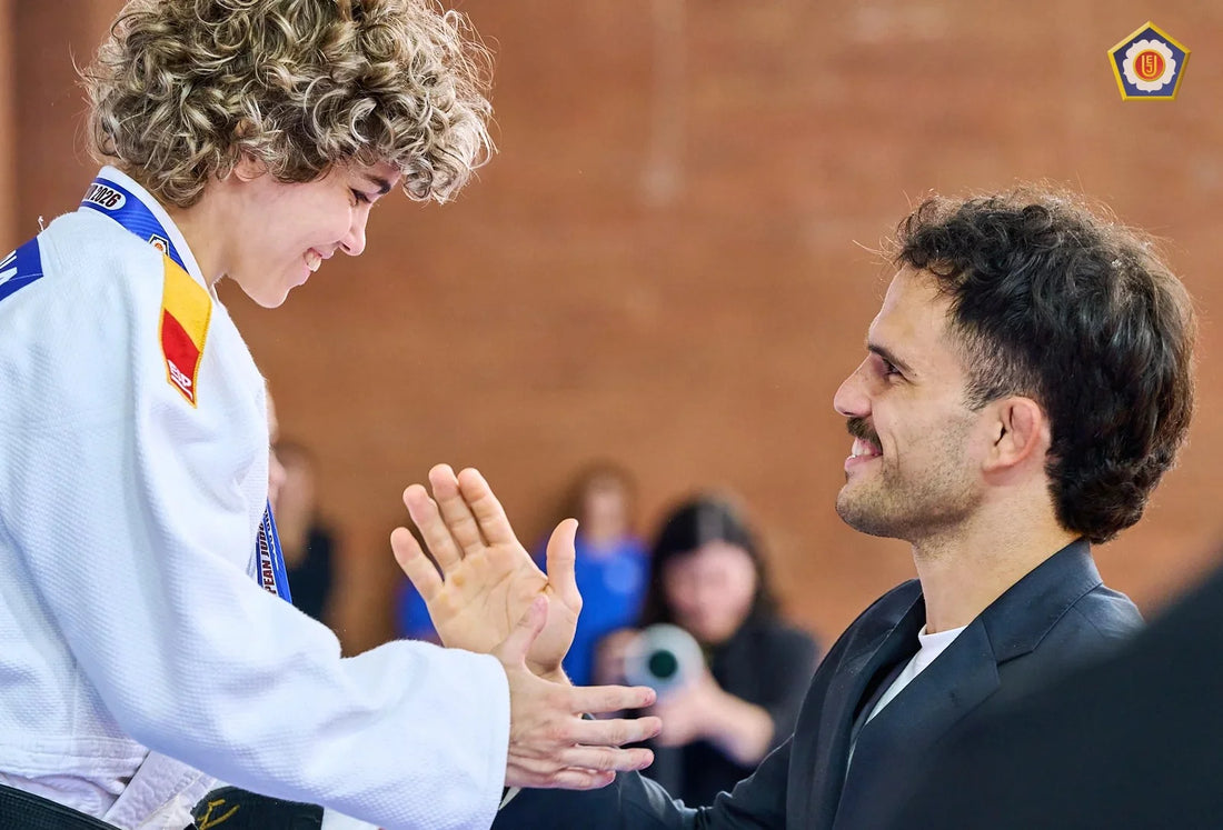 A judoka in a white gi shakes hands with a smiling man in a dark jacket as spectators watch behind them.