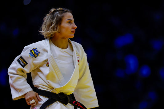 Female judoka in a white judogi and black belt standing in profile against a dark background with blue lights.