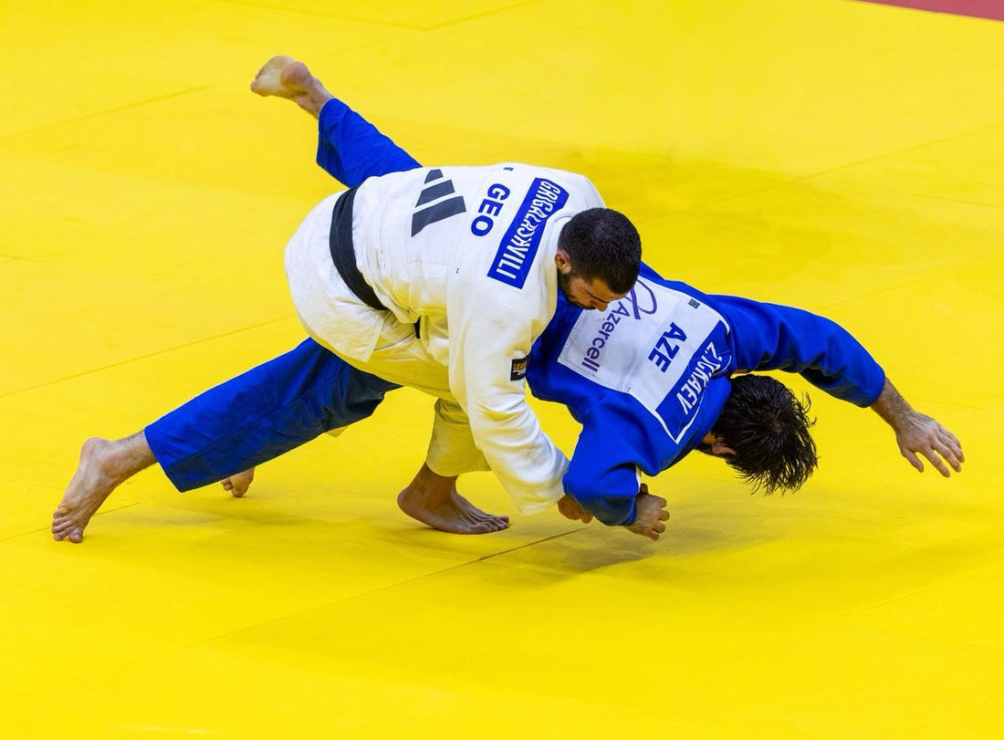A judoka in a white gi throws or controls a judoka in a blue gi on a bright yellow tatami.