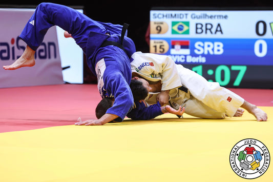 Boris Rutovic in a white judogi battles Guilherme Schimidt in blue during the -90 kg final in Budapest, with the BRA-SRB scoreboard behind.