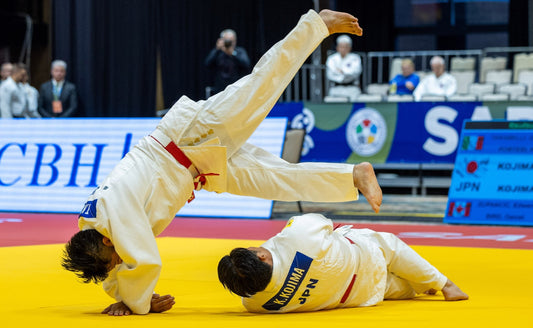 Two judoka in white judogi on a yellow mat, with one athlete lifted upside down while the other lies on the mat.