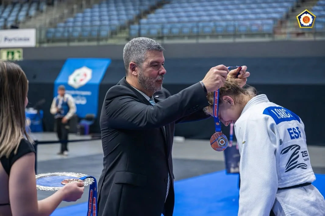 A man in a dark suit places a medal over the head of a judoka in a white judogi on a blue mat.