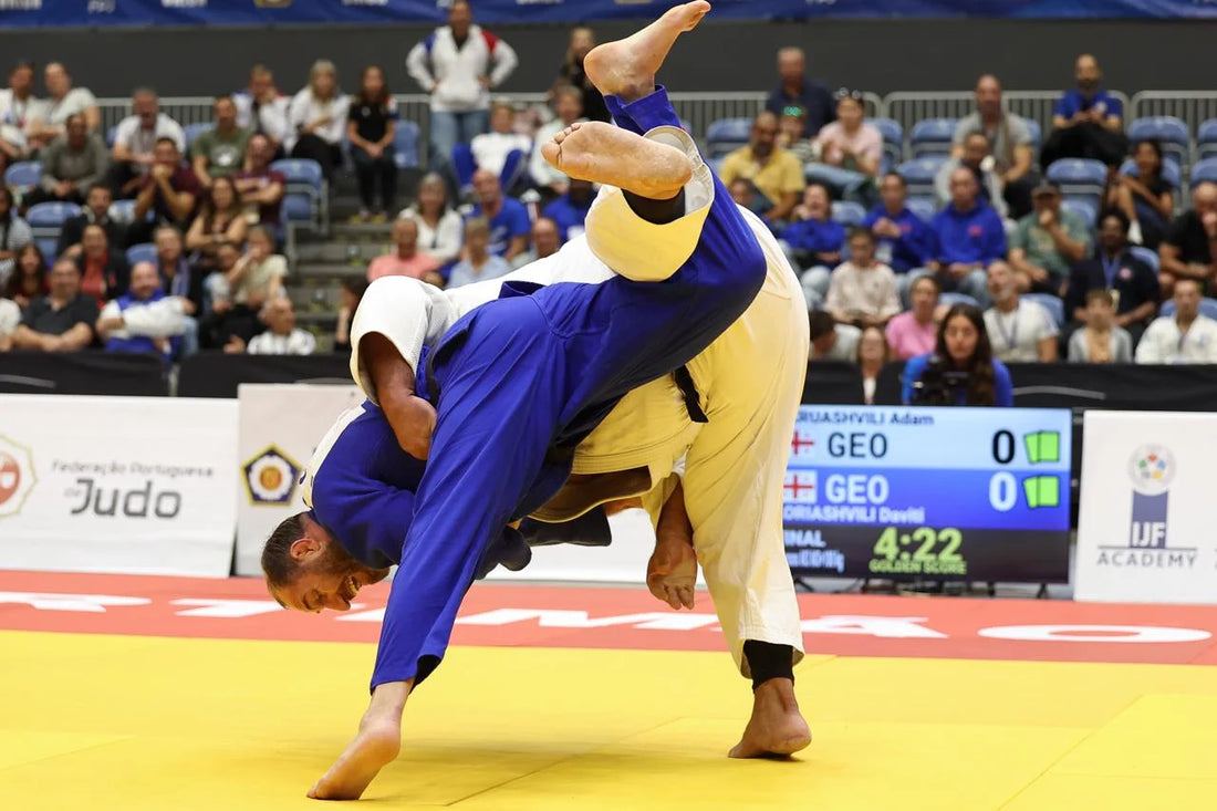 A judoka in a blue judogi is lifted upside down by an opponent in a white judogi on the yellow tatami, with spectators behind them.